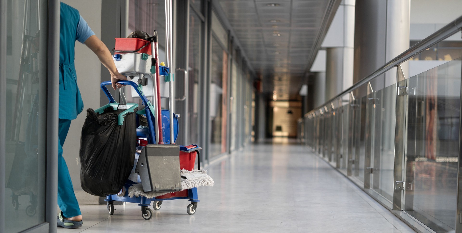 Ein Mitarbeiter zieht einen Trolley für die Reinigung von Büros. Die Putzfrau ist mit der Arbeit beschäftigt.