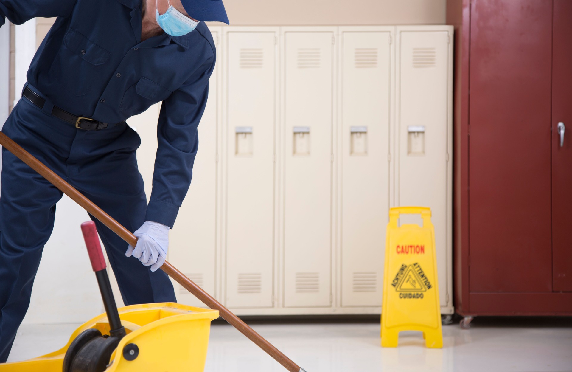 Senior Adult Janitor mops Boden in Bürogebäude.  Er trägt eine schützende Gesichtsmaske.